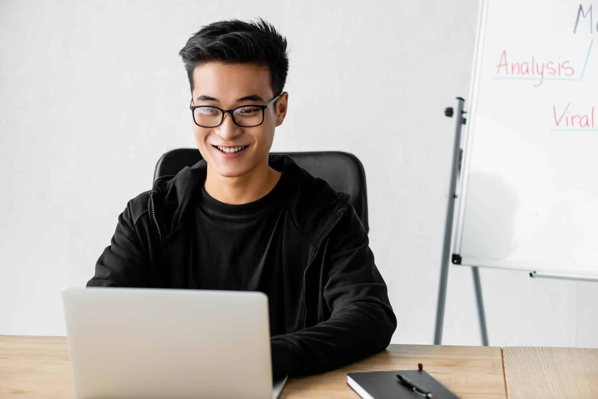 smiling seo manager in glasses sitting at table and using laptop